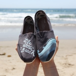 Custom, hand painted Life is Better at the Beach TOMS shoes featuring lettering and a gradient wave pattern. Displayed at Santa Monica Beach in California.
