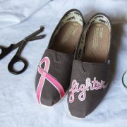 Custom, hand painted Breast Cancer Fighter grey TOMS shoes featuring the word "fighter" and the pink breast cancer awareness ribbon. Flatlay with medical tools in hospital background.